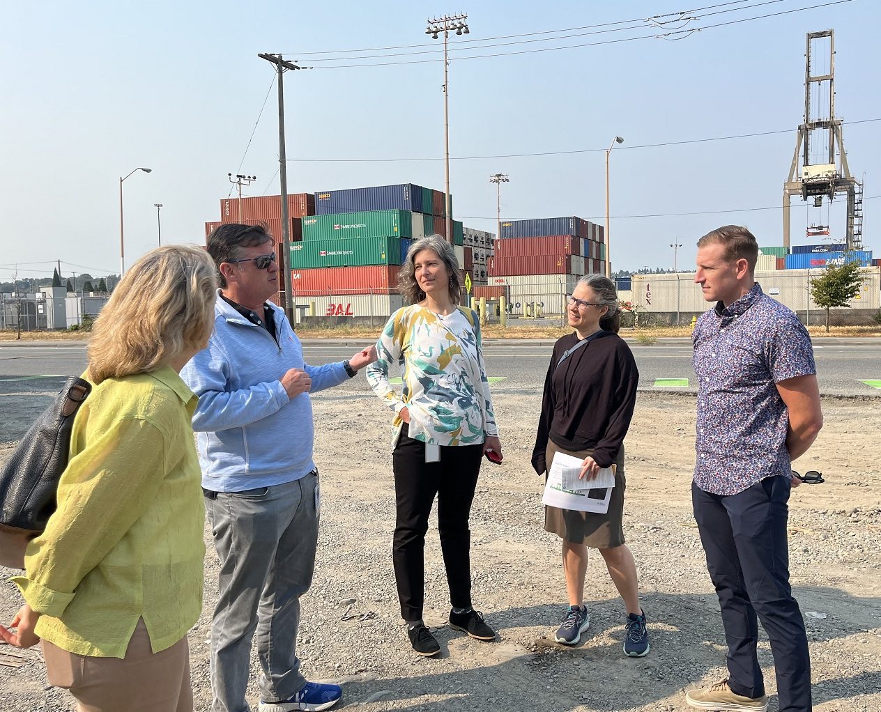SDOT Director Greg Spotts tours East Marginal Way with the SDOT project team and representatives from the Port of Seattle. Five people stand talking in the foreground, with large Port of Seattle cargo containers and a crane in the background on a sunny day.