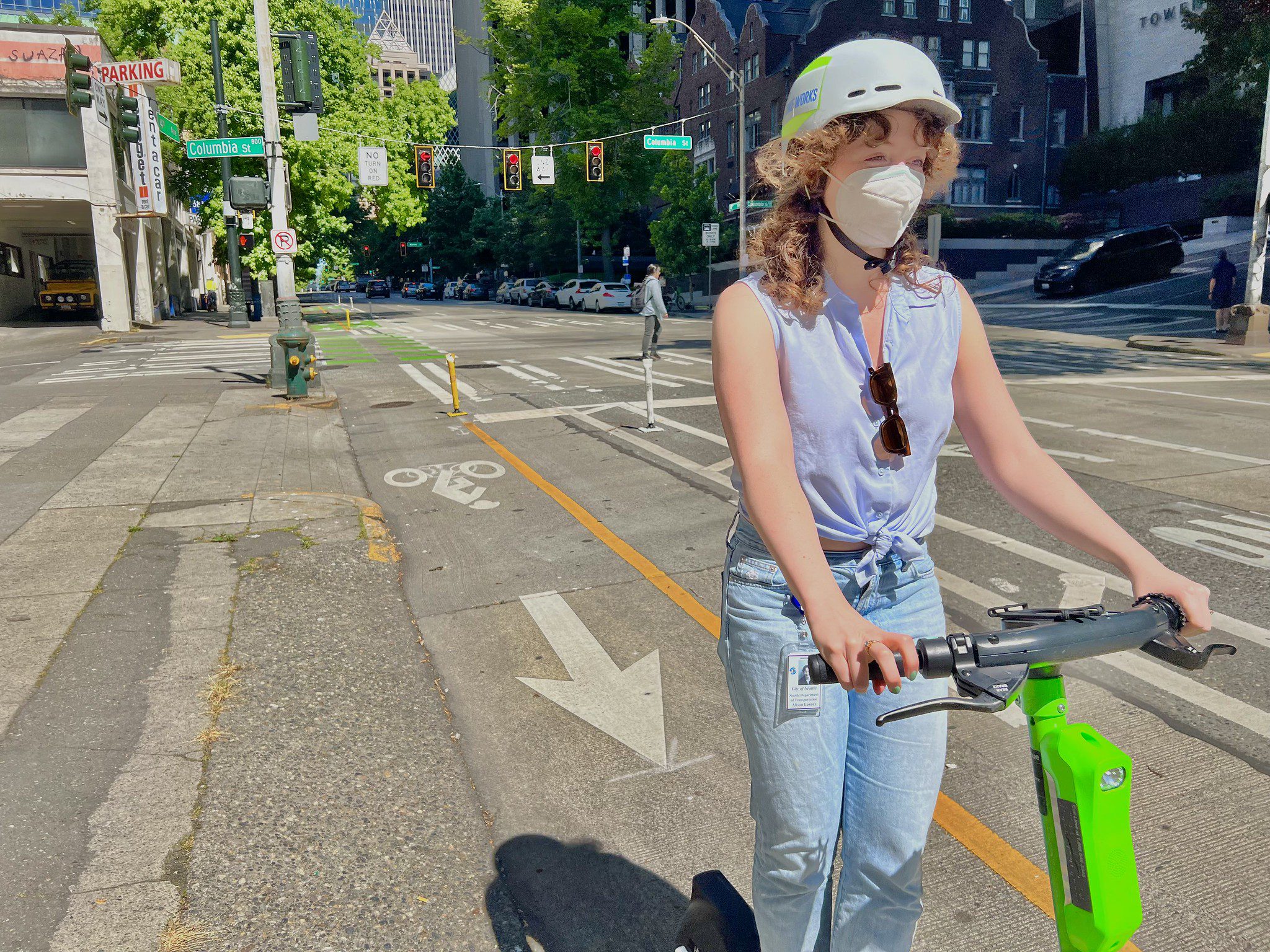 A scooter share rider travels along a protected bike lane on 4th Ave in downtown Seattle. The rider is wearing a white helmet and a mask, riding a green scooter down the protected bike lane.