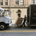 A person unloads large cardboard boxes and packages from a truck in a center lane in Seattle. Two trucks and a large building are in the background, with a painted yellow line in the foreground.