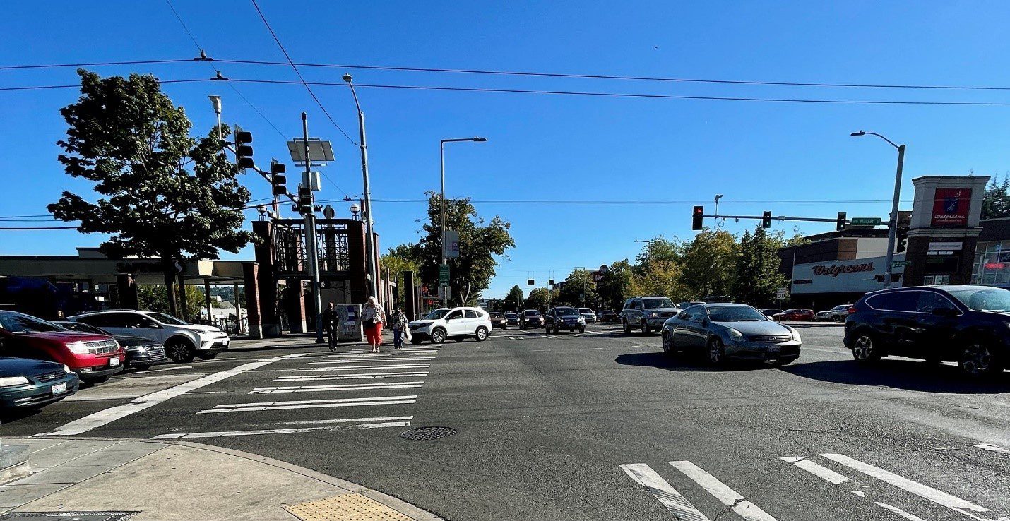 People crossing the street at 15th Ave NW and NW Market St. Marked crosswalks, cars, and blue skies are in the foreground and background.