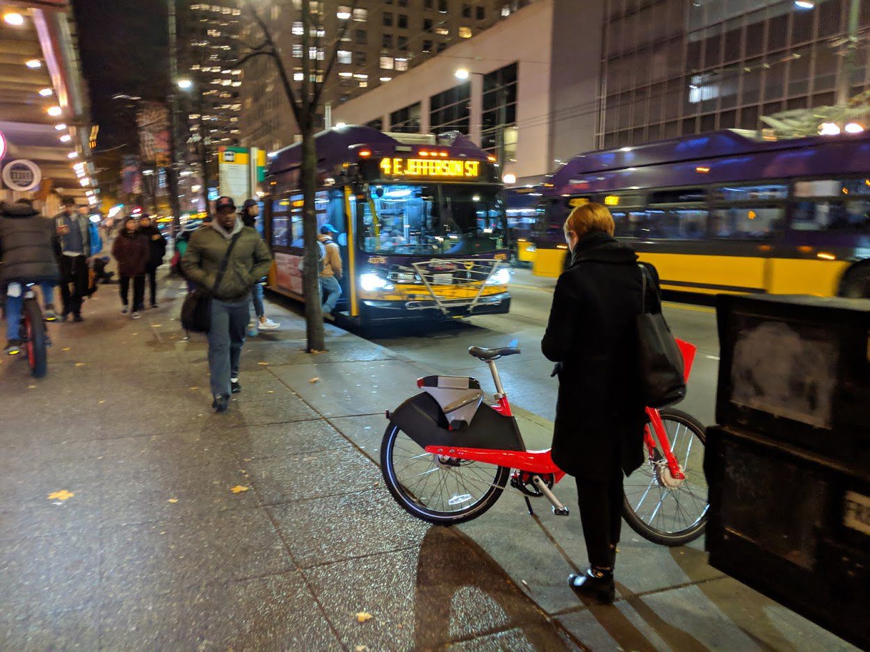 People travel along 3rd Ave in downtown Seattle at night. People walk along the sidewalk as buses pass by in the street. A bike is in the foreground, with large buildings in the background.