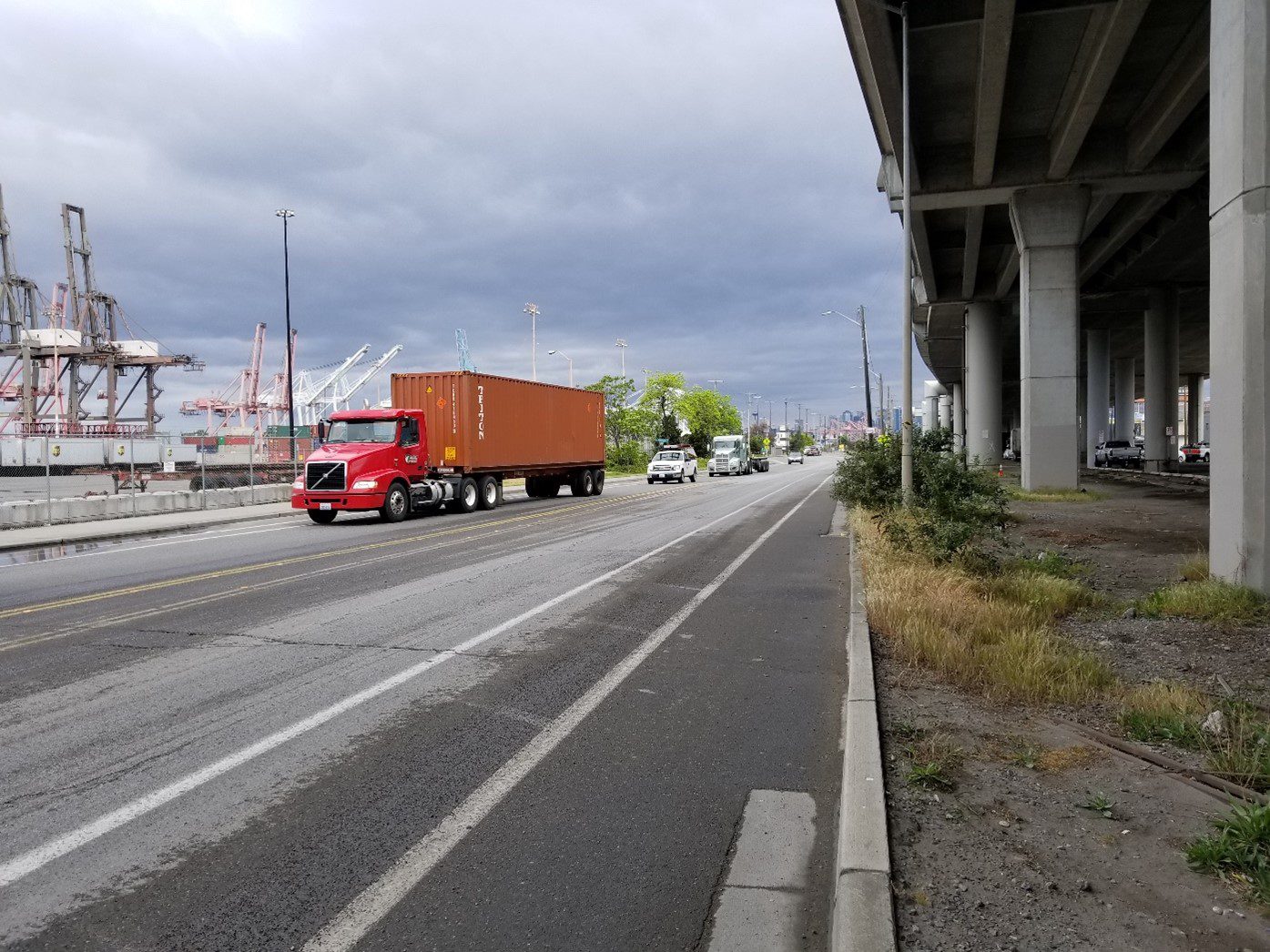 Photo of a large freight truck traveling along East Marginal Way S, with vehicles behind it. Large Port of Seattle cranes are to the left, and the street in the middle. An elevated roadway is to the right, with gray columns.