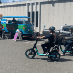 People try out riding bike share equipment at a public demonstration and training event. A blue van and large building are in the background, with paved parking areas in the foreground.