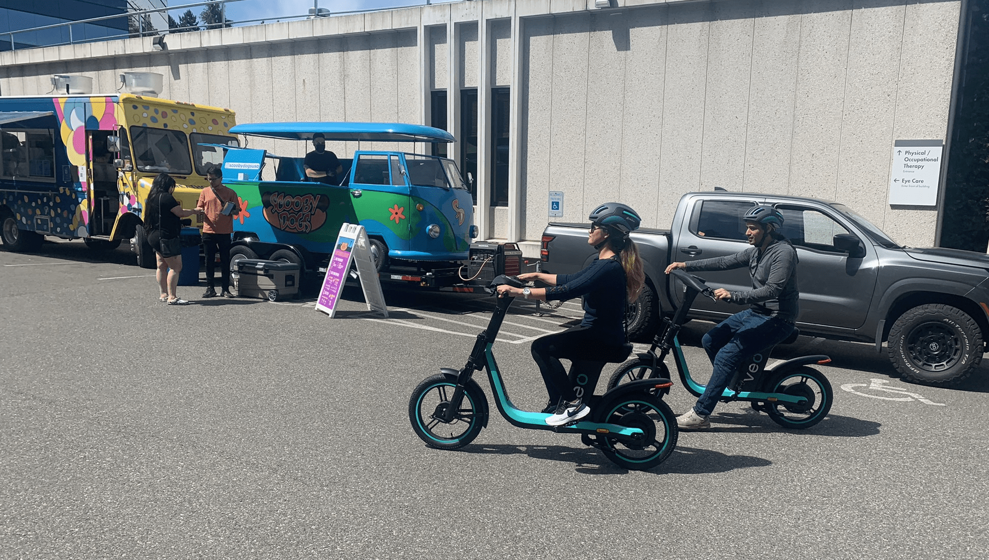 People try out riding bike share equipment at a public demonstration and training event. A blue van and large building are in the background, with paved parking areas in the foreground.