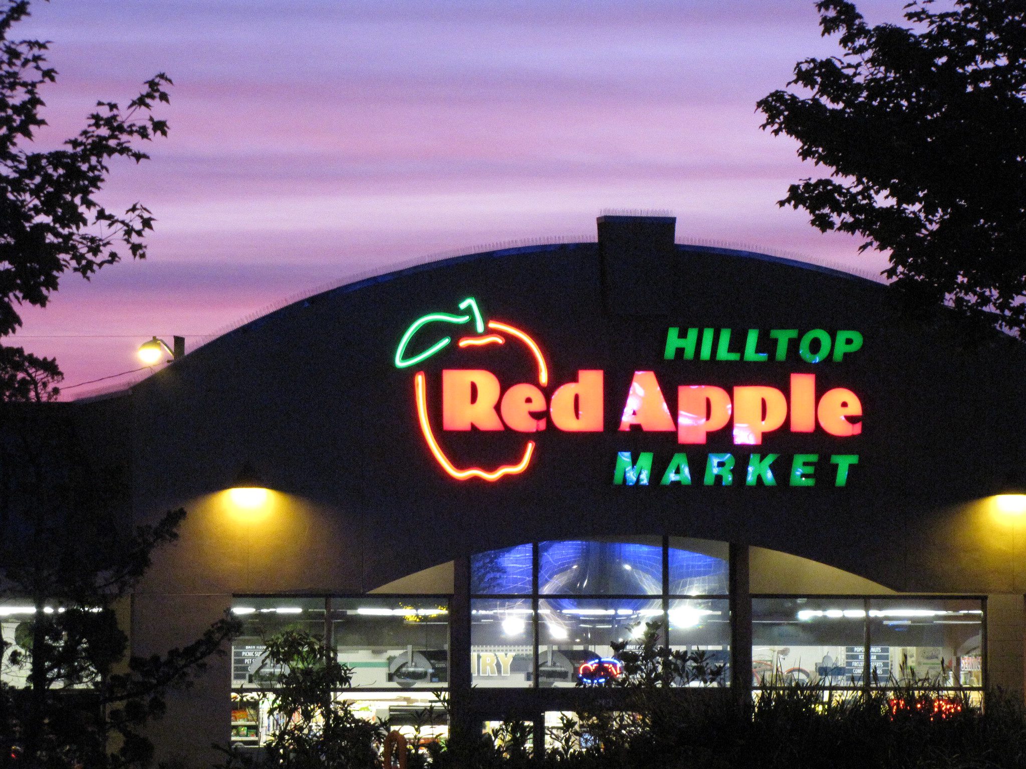 Photo of the Hilltop Red Apple Market on Beacon Hill in the late evening. A large red apple neon light illuminates the lettering of the grocery store, with views inside as it gets dark out.