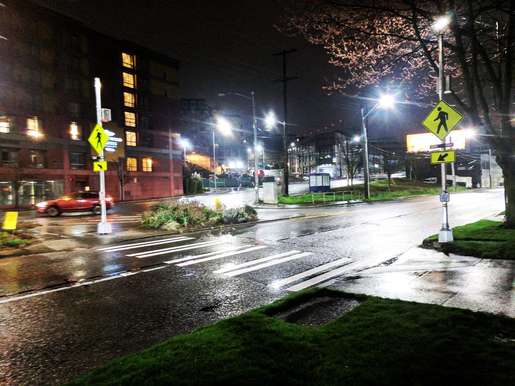 An intersection at night. Rapidly flashing beacons alert drivers to the potential for pedestrians. A red car passes through the crosswalk. Large buildings are in the background, with grass and landscaping in the foreground.