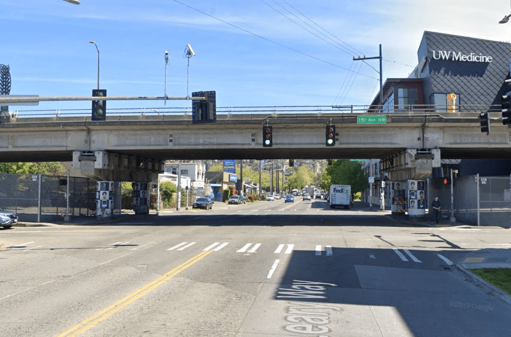 View of the Leary Way Bridge. The bridge spans above Leary Way NW. A traffic signal, street sign, street, and cars are in the rest of the image, with a large building in the background showing a UW Medicine sign.