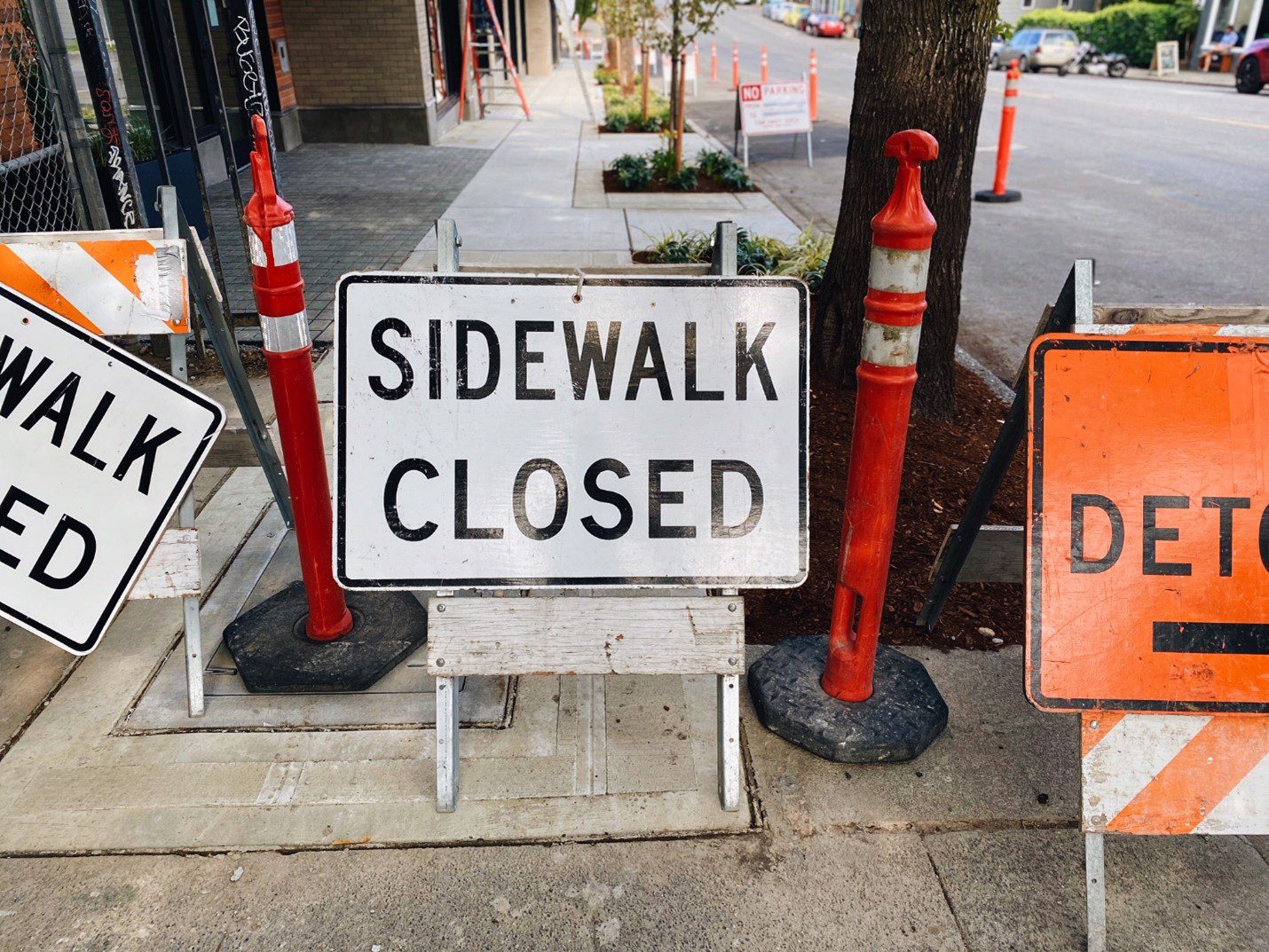 A large white sign indicates that a sidewalk is closed ahead. Next to it are several orange signs and orange vertical construction markers.