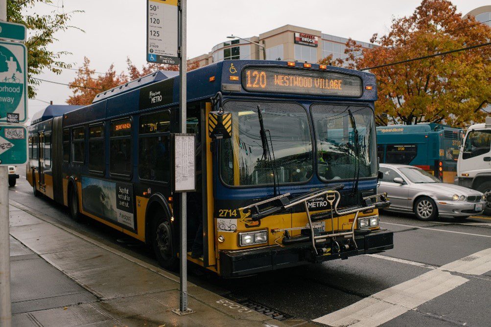 A King County Metro bus route 120 at a bus stop in Seattle. Large trees and other vehicles are also in the photo, along with the bus stop sign.