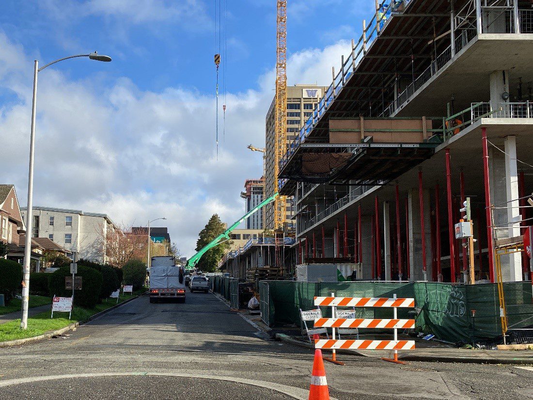 A large building under construction in the University District of Seattle. A large orange barricade and smaller sidewalk closed signs are next to the project site fencing. Large trucks and equipment are in the background, with homes and buildings to the left side.