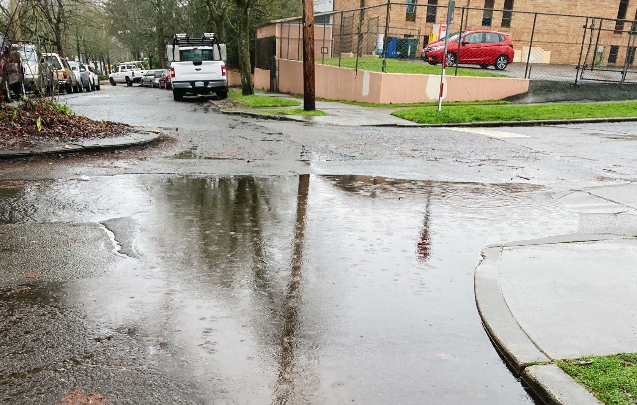 Example of water pooling on a street in Seattle near a clogged storm drain. The street and sidewalk are in the foreground, with a car and truck, and building in the background.