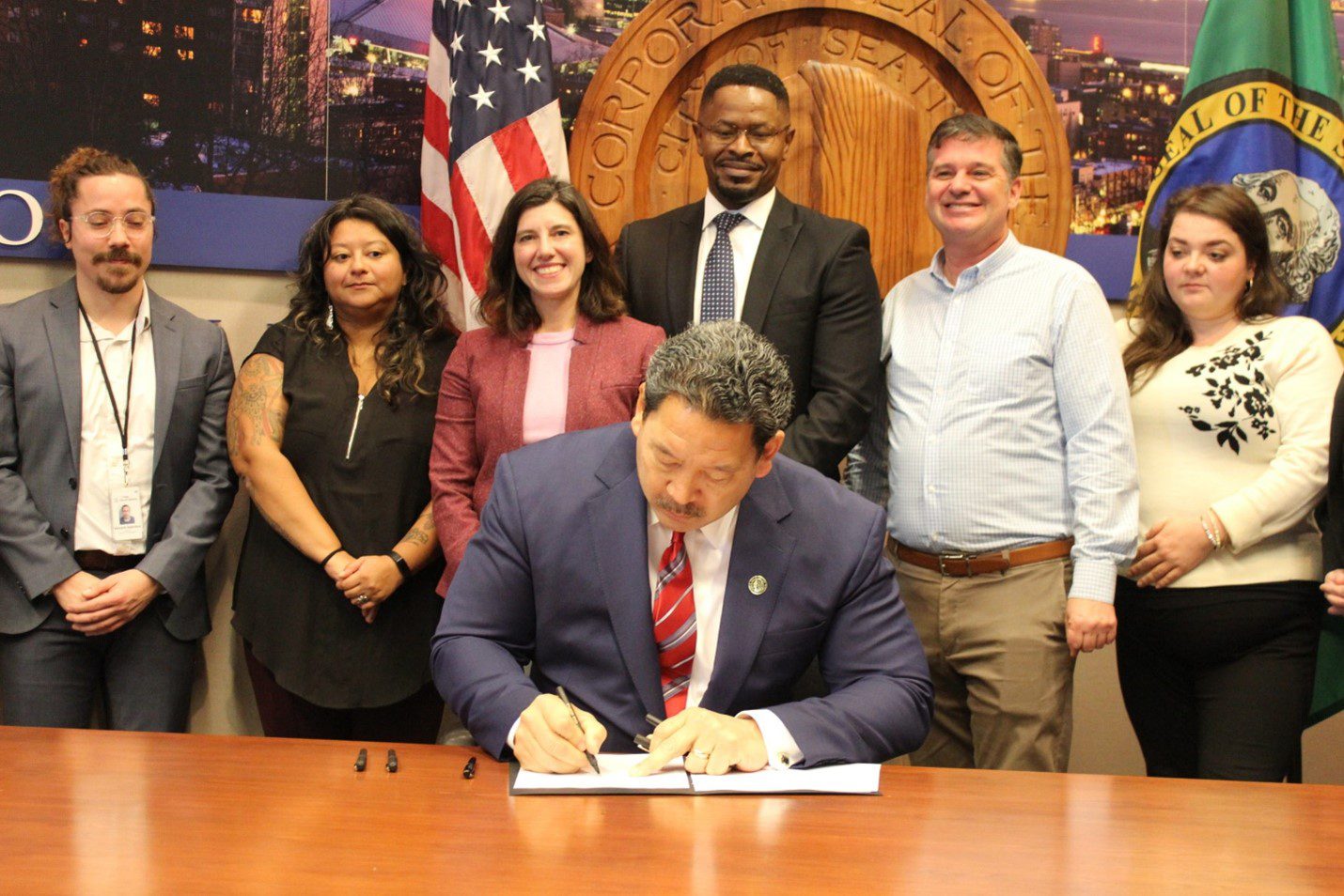 Photo of Seattle Mayor Bruce Harrell signing a new executive order. Several City leaders and department heads stand behind the Mayor, while watching him sign the order. The American flag, Washington State flag, and City of Seattle seal are in the background.