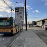 A bus travels away from the camera on a sunny day. A white sign notes curb lane, buses only, from 7-9AM and 3-7PM on weekdays. Sidewalk and buildings and parked cars are in the background.