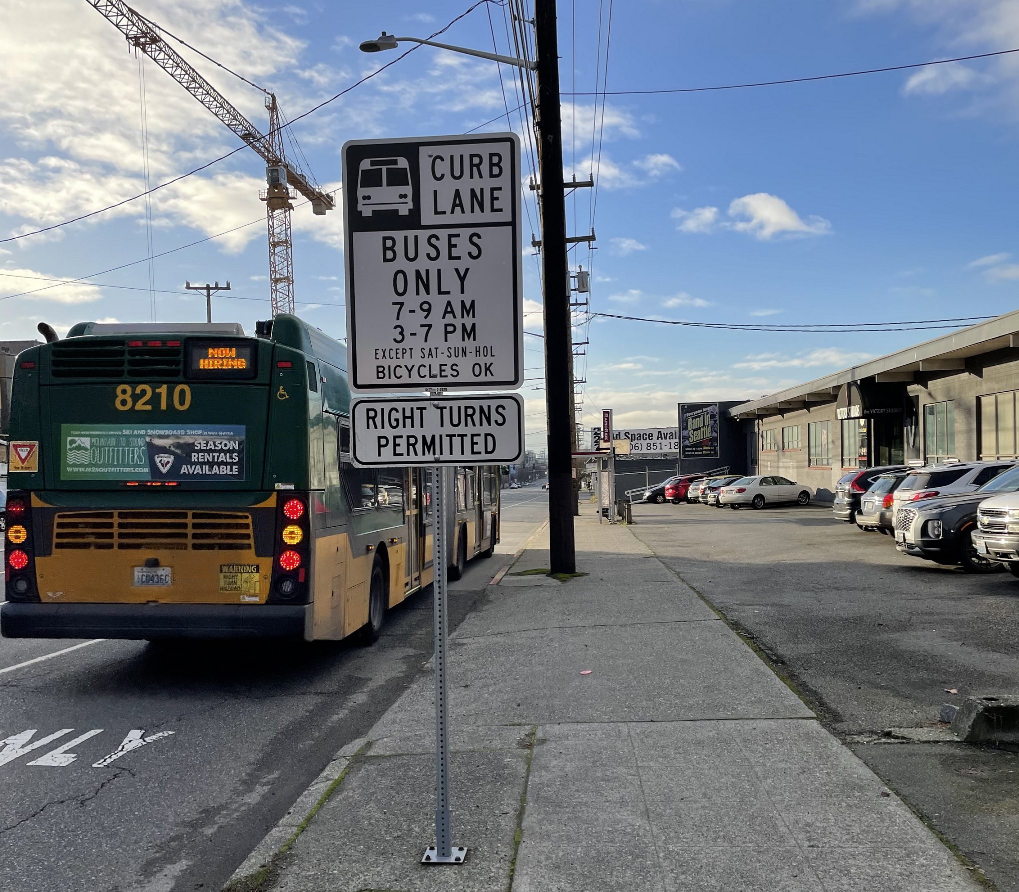 A bus travels away from the camera on a sunny day. A white sign notes curb lane, buses only, from 7-9AM and 3-7PM on weekdays. Sidewalk and buildings and parked cars are in the background.