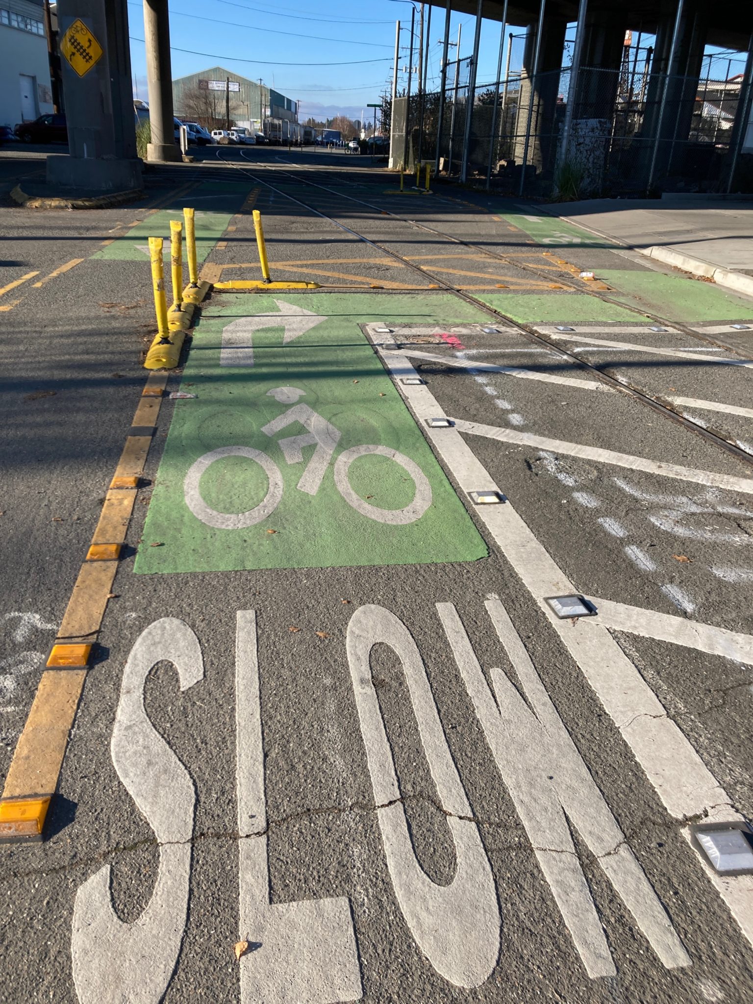 Picture of current layout of bike lanes under the Ballard Bridge. Green pavement markings and yellow posts indicate that bike riders should turn right at a 90 degree angle to cross train tracks.