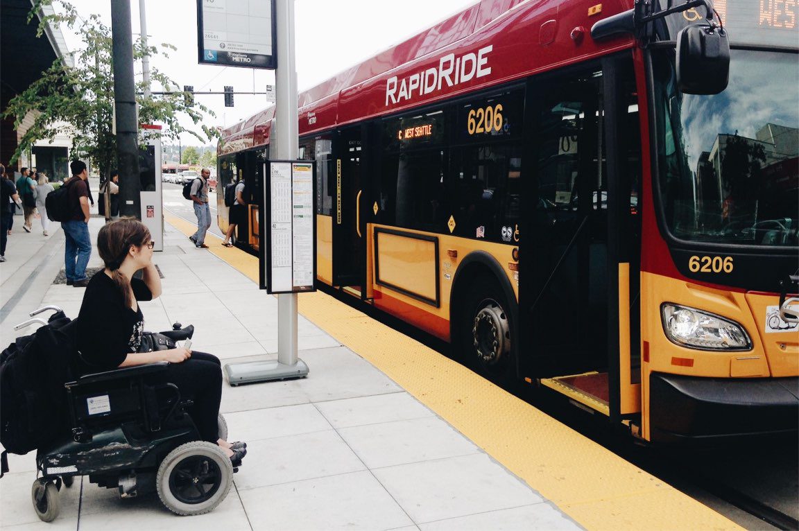 A person in a power wheelchair prepared to board the RapidRide C Line bus to West Seattle. The bus is on the right and people also board the bus in its rear doors, while all doors are open.
