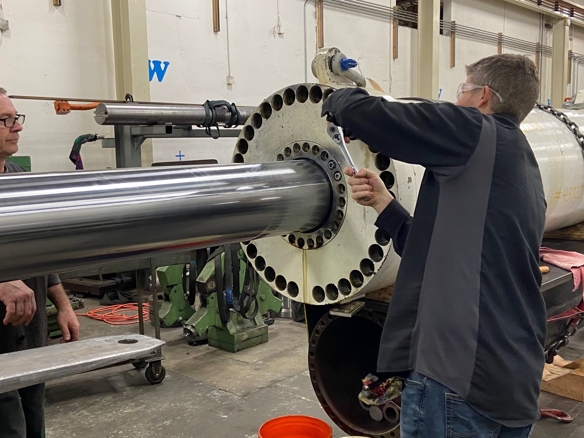 Specialists working on the massive hydraulic turning cylinder. The person working wears safety glasses and a black jacket, inside a building with white walls.