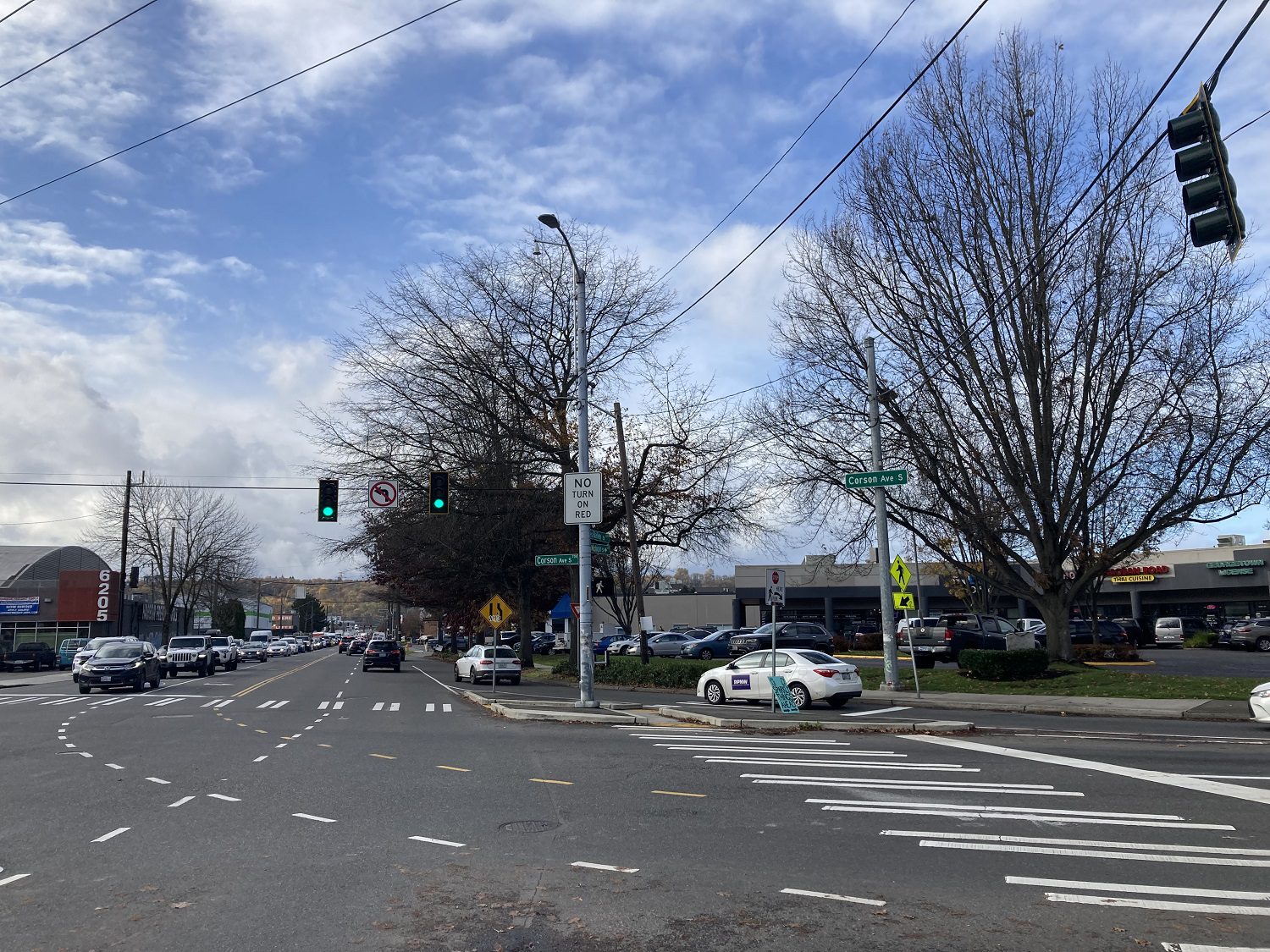 Photo of an intersection with cars visible and large trees on a clear day. A marked crosswalk is on the right side of the photo.