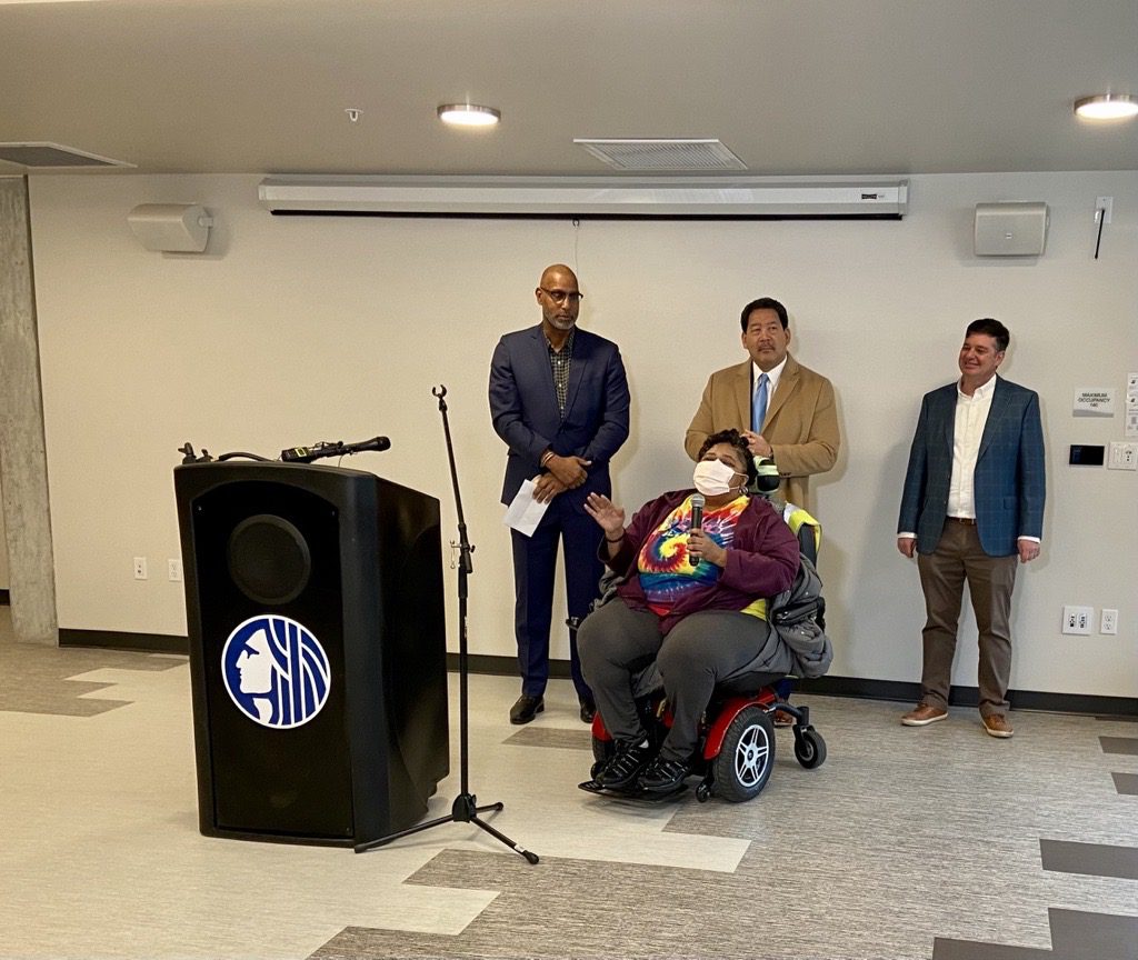 A woman in a power wheelchair wearing a mask gives remarks at a media event, next to a podium. Three men stand behind her, listening.