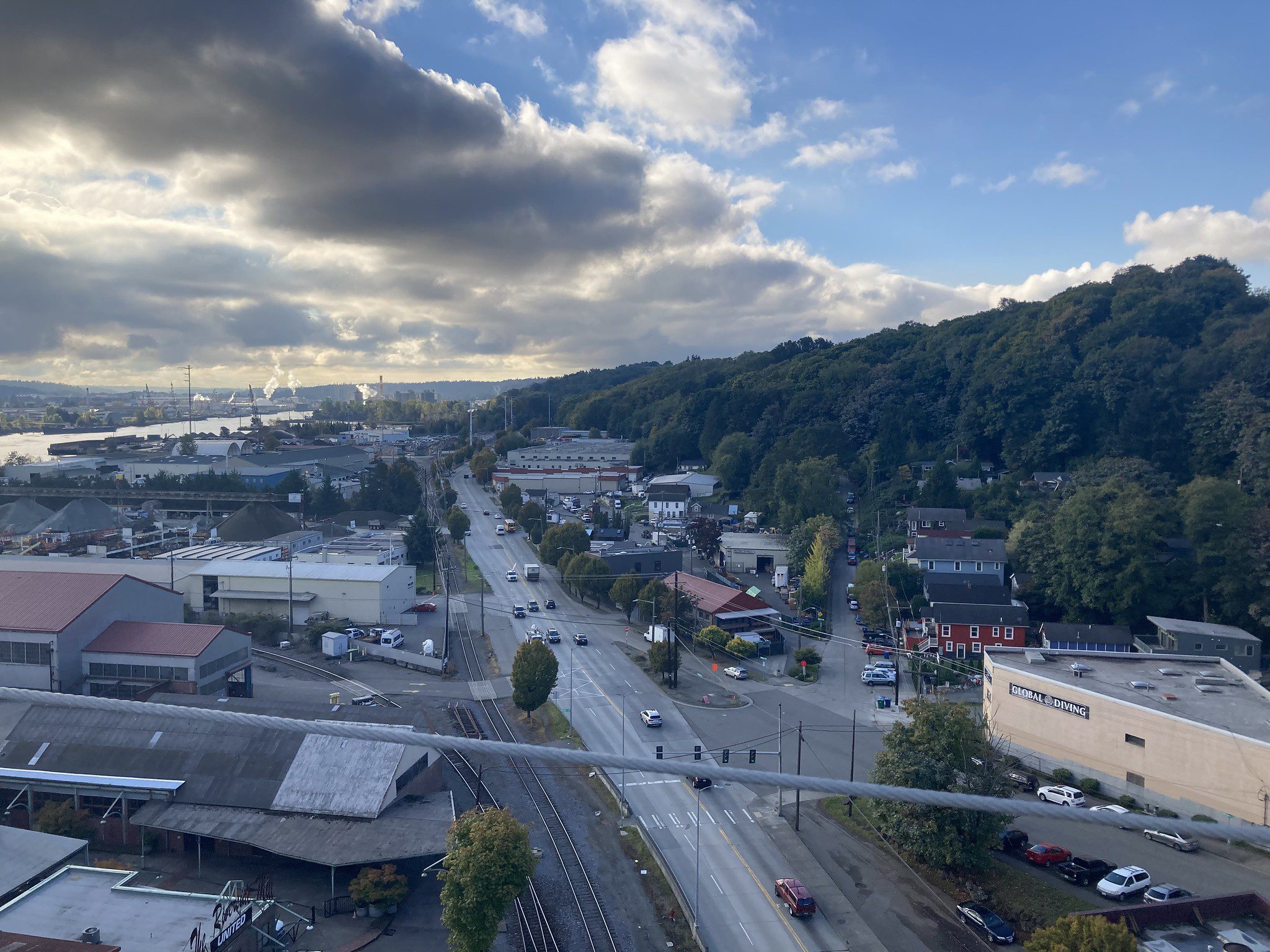 Photo from a high vantage point looking out at a street with buildings, cars, and traffic lights. Bright blue skies and clouds are above, with a hillside to the right.