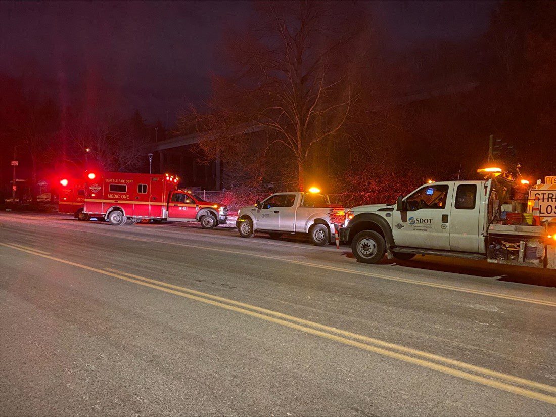 Two large white trucks and two red ambulances respond to a scene at Airport Way at night. Large trees are in the background and pavement is in the foreground.