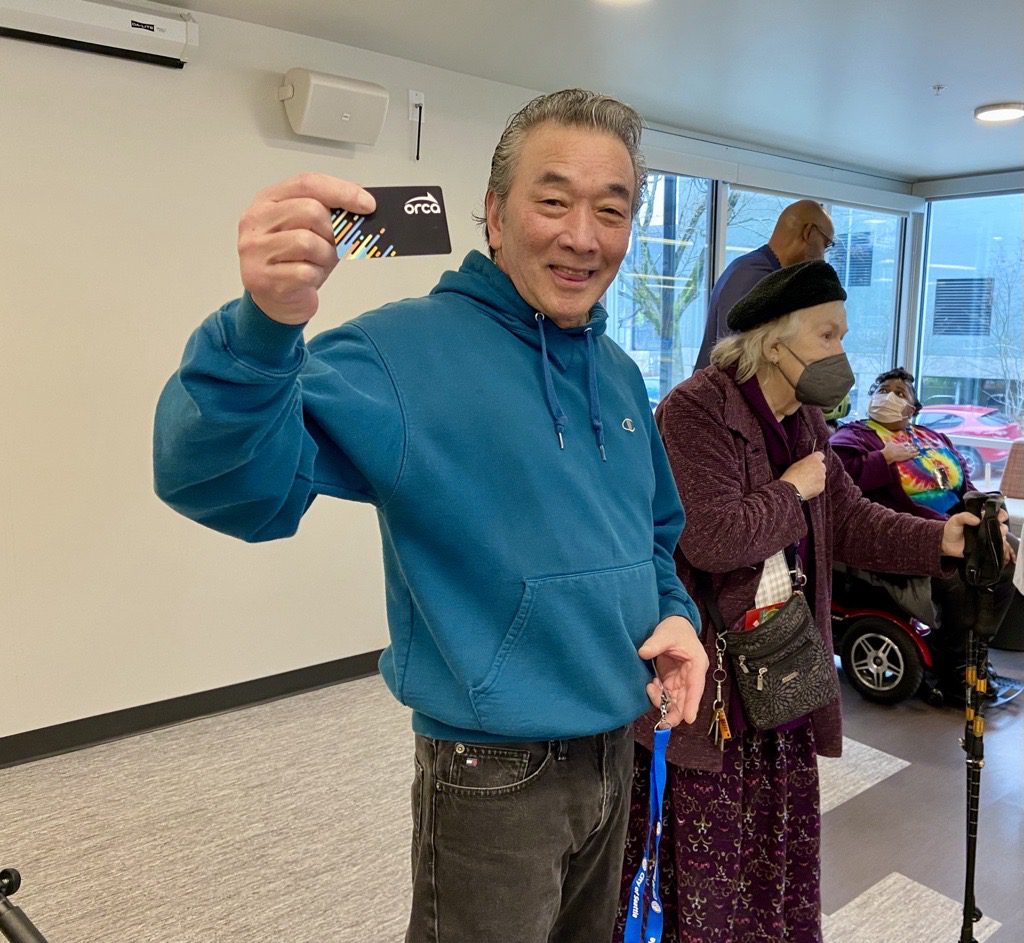 A man wearing a blue sweatshirt and jeans holds up a black ORCA card while smiling at the camera. A woman wearing a mask stands next to him, with two other people in the background inside a building.
