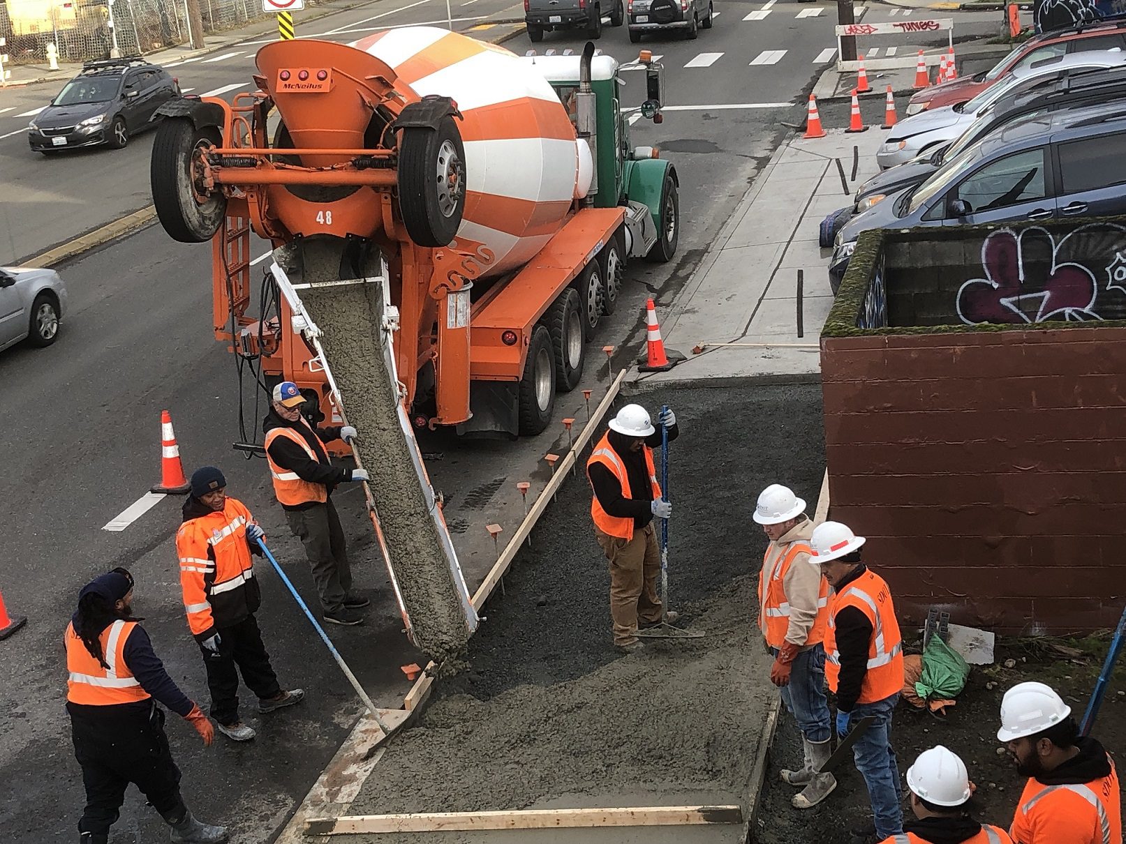 Eight people wearing orange vests work to pour concrete on a section of sidewalk next to the street. A large cement mixer truck is in the upper left, and a worker spreads the mix out for placement. Parked cars and orange cones are also in the picture.