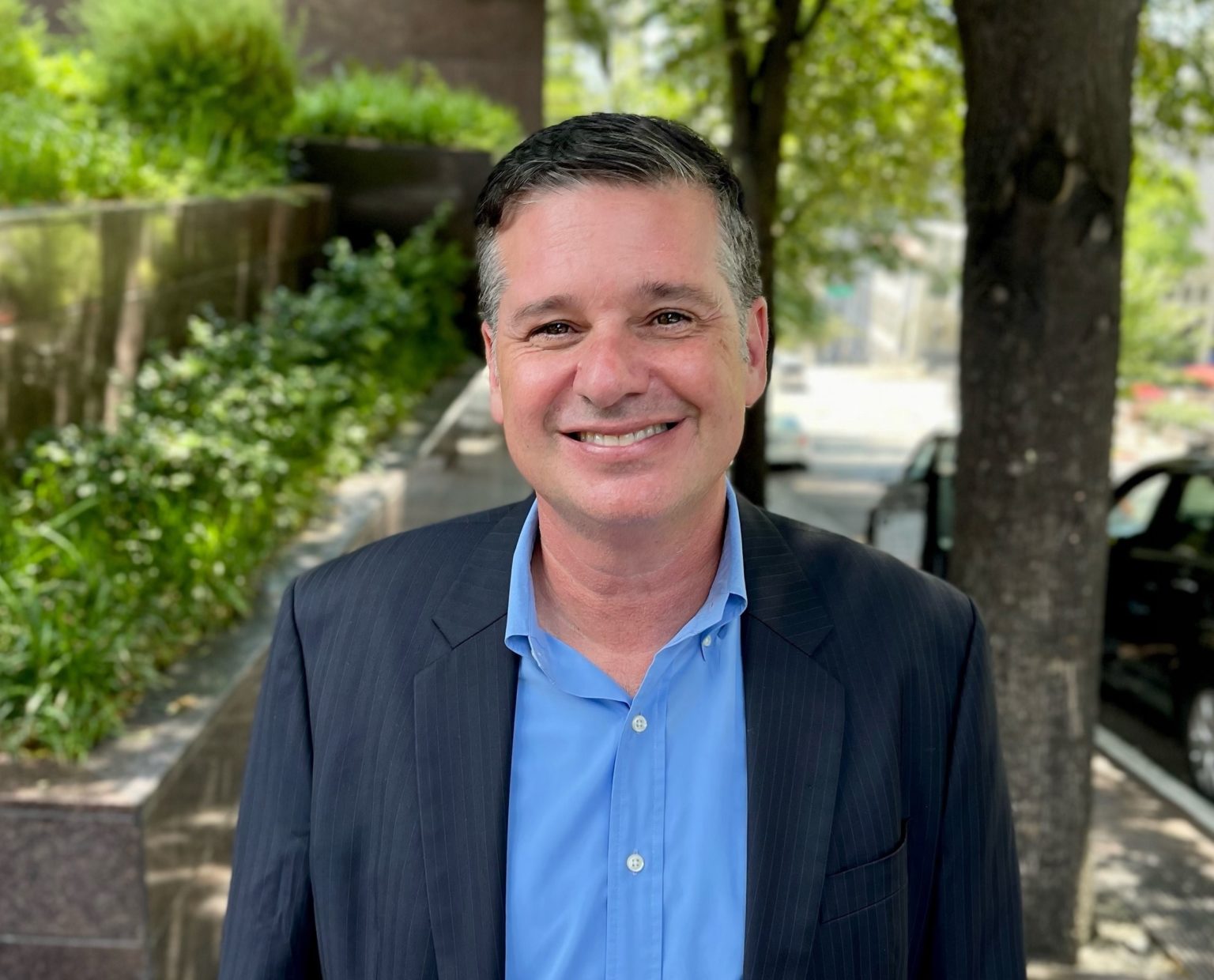 Photo of SDOT Director Greg Spotts smiling at the camera on a sunny day. Bushes and trees, as well as parked cars, are in the background. Greg is wearing a navy blue suit jacket and a blue dress shirt.