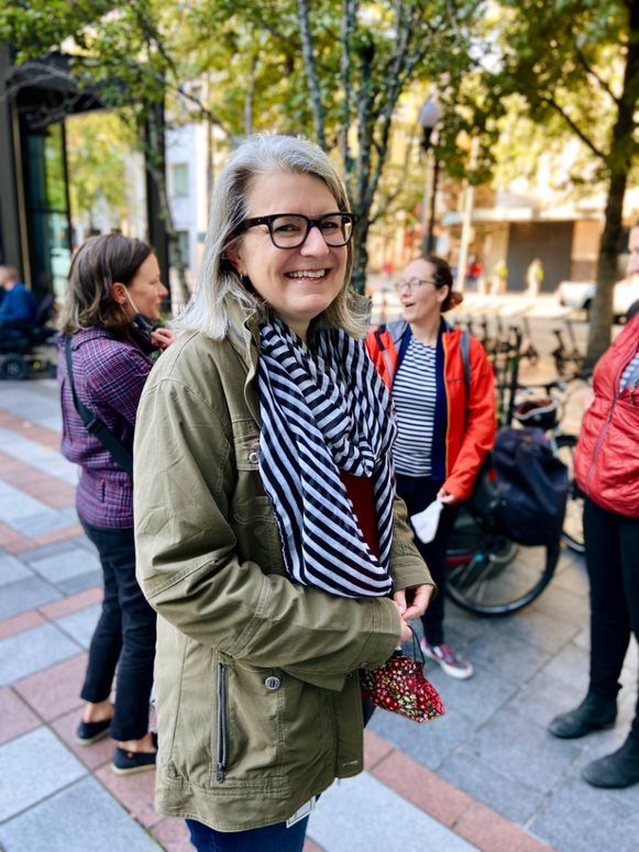 SDOT Senior Deputy Director Kristen Simpson smiles at the camera in a photo from downtown Seattle. Other people are in the background, as well as tiles and large trees.