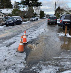 Two orange cones mark a large pothole near snow and ice on a street, with parked cars in the background.