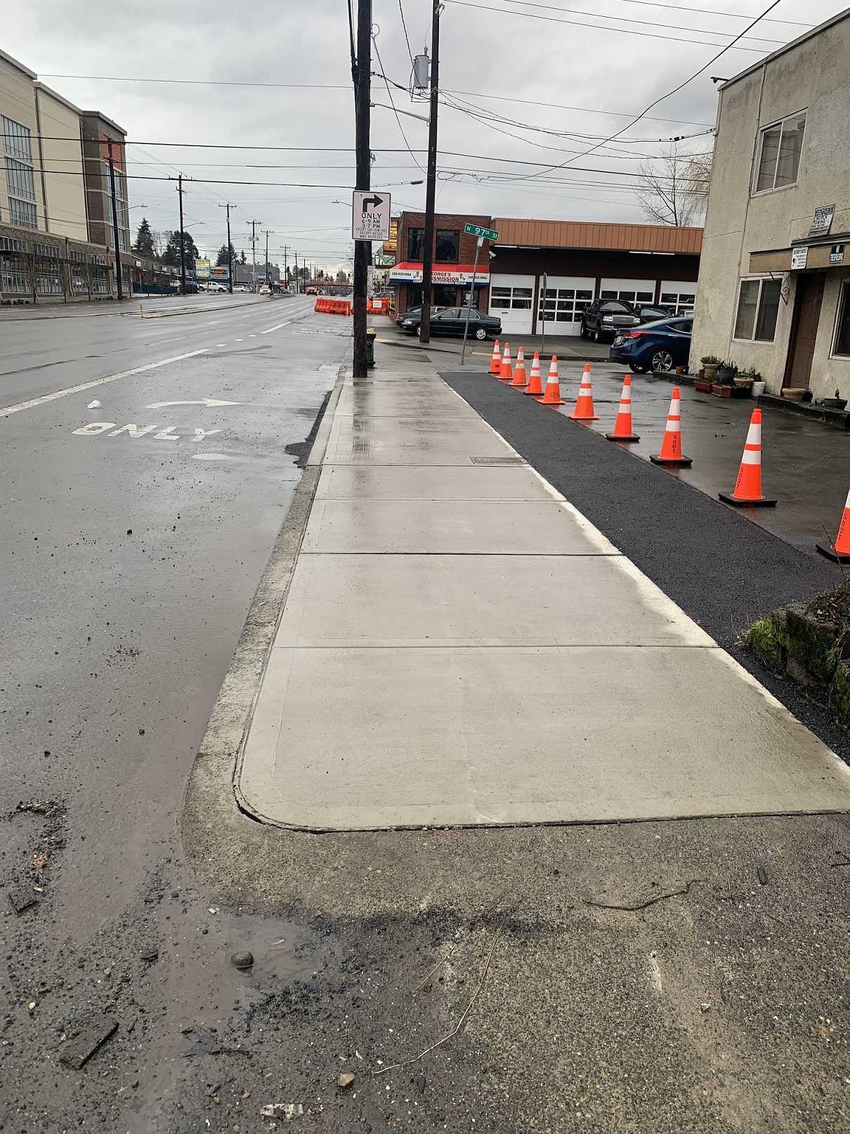 A smooth section of sidewalk that is wet is in the middle of the image. Orange cones are to the right. Parked cars and a street are in the background, as well as several buildings.