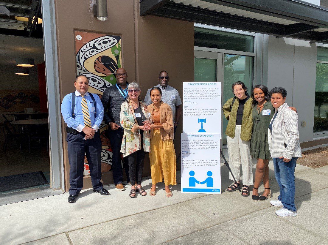 Photo of staff and community members standing together to celebrate the publication of the Transportation Equity Framework in summer 2022. Eight people stand near a building and a large display board, on a sunny day.