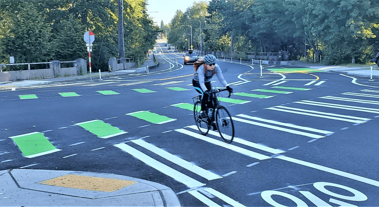 Photo of a person biking on a newly repaved 15th Ave NE. The person is turning into a protected bike lane, with large trees in the background and curb ramp in the foreground.