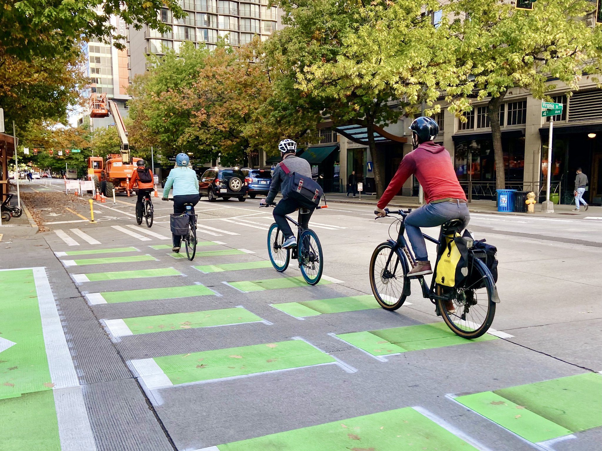 People bike along 4th Ave in downtown Seattle and the Belltown neighborhood, on a sunny day. Large trees are in the background, and painted green protected bike lane in the foreground.