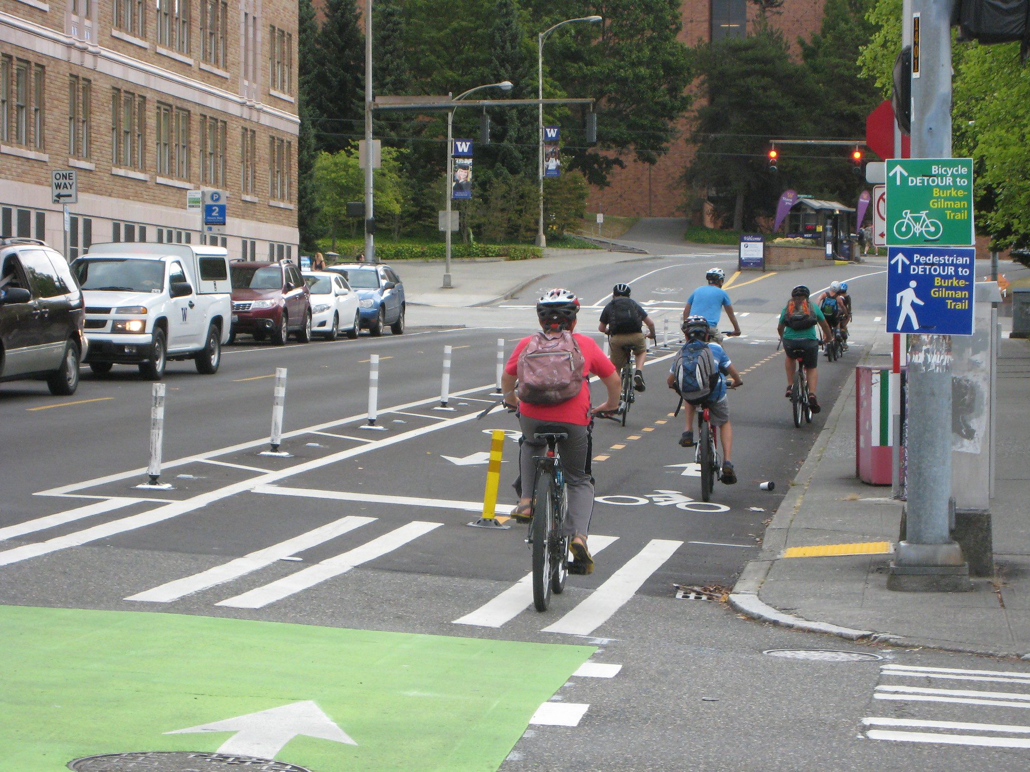 Several people including young people bike along a protected bike lane. Large buildings and parked cars are in the background, as well as utility poles and signs.