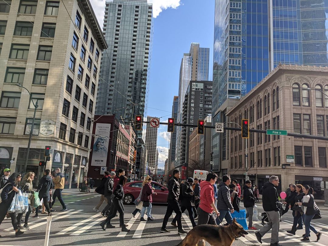 Many people cross the street in downtown Seattle, along Pine Street. Large towers and buildings are visible in the background. A street sign says Pine St.