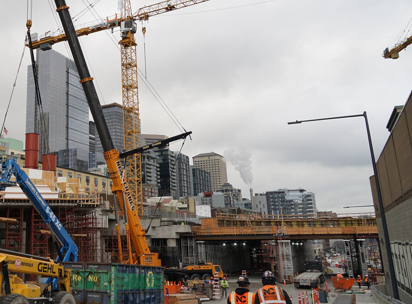 Construction workers on a construction site on a cloudy day. A large crane and temporary bridge false work are in the background, as well as several large buildings.