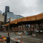 Photo of a bridge under construction. Large buildings are in the background with the street in the foreground, on a cloudy day.