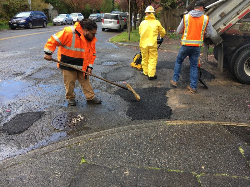 Three people wearing bright orange and yellow safety gear work to fill potholes in a street. Parked cars are in the background, as well as a sidewalk in the foreground.