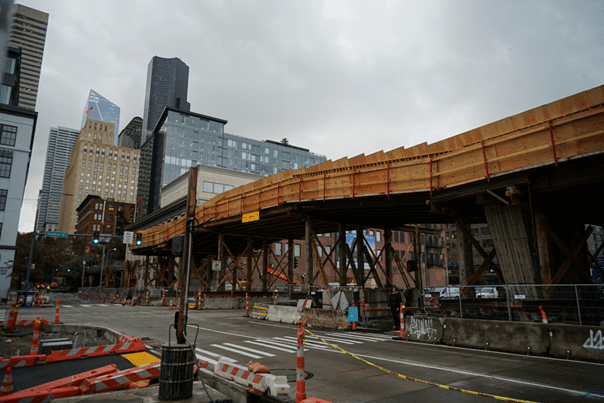Photo of a bridge under construction. Large buildings are in the background with the street in the foreground, on a cloudy day.