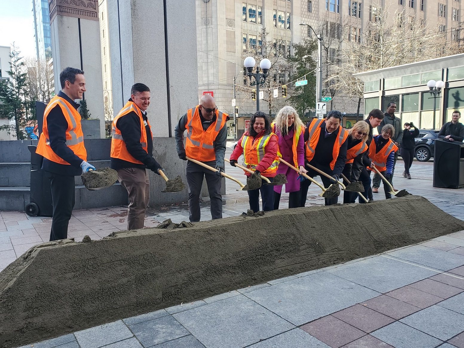 Several people wearing orange vests shovel dirt in the city, along a plaza area, with large buildings in the background.