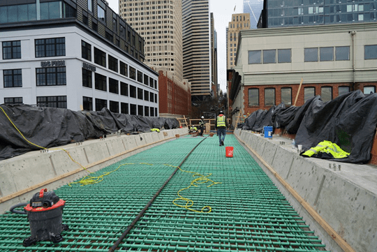 Construction workers stand on green steel rebar, at a construction site. Large buildings are in the background. A yellow cable runs along the green rebar.