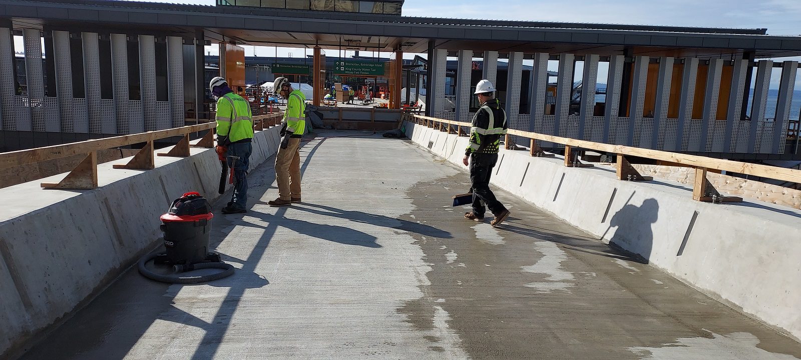 Three construction workers wearing safety vests and hardhats stand atop a concrete bridge next to a large entry building, on a sunny day.