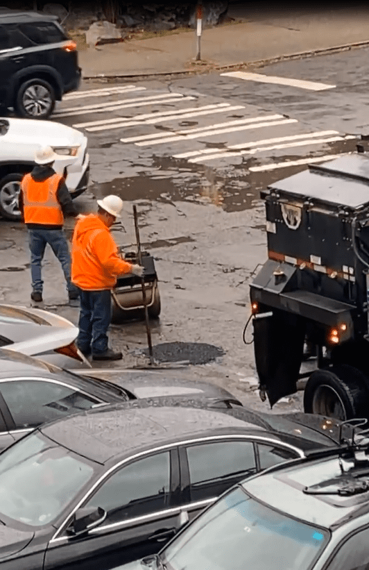 Photo of two people wearing bright orange construction gear and white hardhats filling potholes in the street next to several parked cars and a work truck.