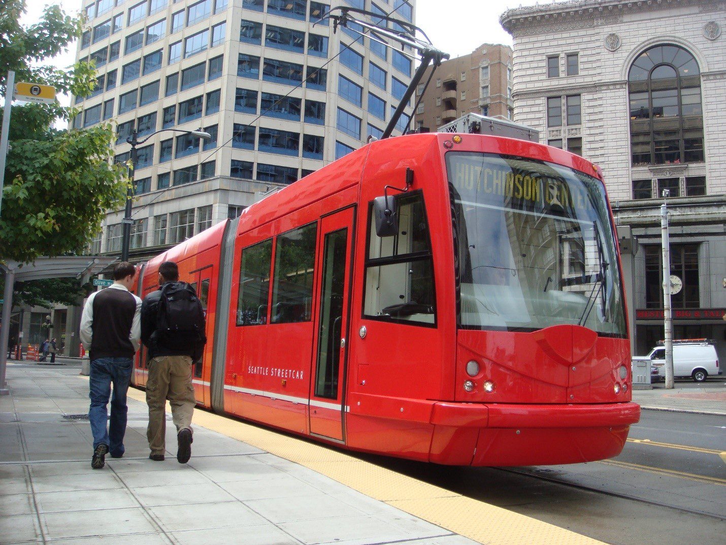 A red Seattle Streetcar train waits for people to board, with large buildings in the background. Two people are to the left.