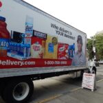 A large delivery truck parked curbside on a cloudy day. Pictures of products and a person are on the side of the truck, as well as a small sandwich board side on the curb.
