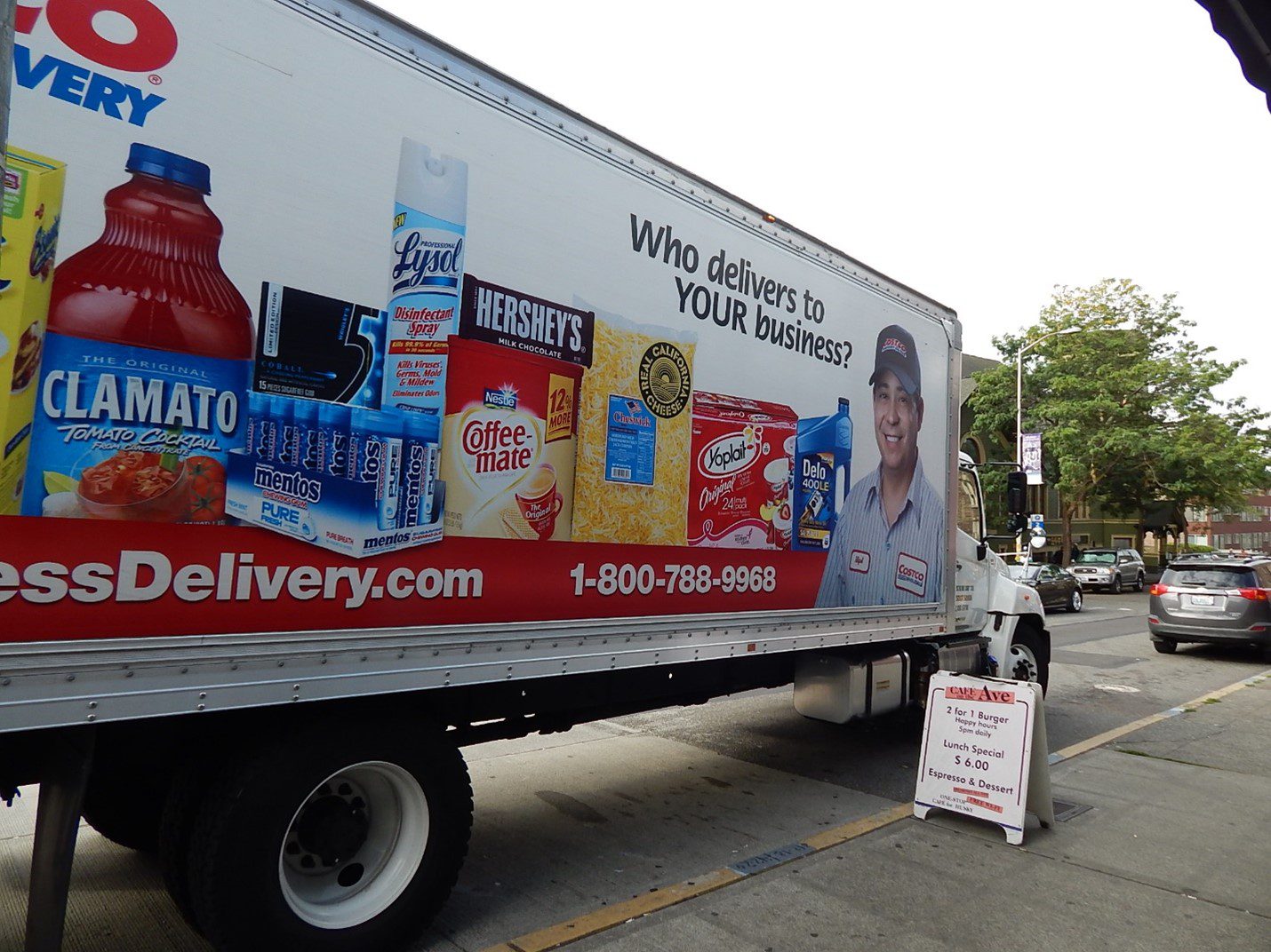 A large delivery truck parked curbside on a cloudy day. Pictures of products and a person are on the side of the truck, as well as a small sandwich board side on the curb.