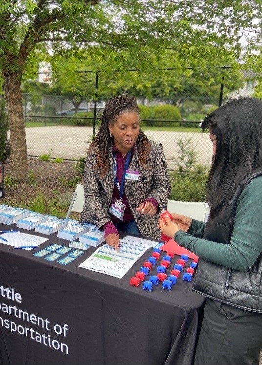 Photo of two women connecting at a community outreach table with giveaways on top of a tablecloth. Trees and a park are in the background.