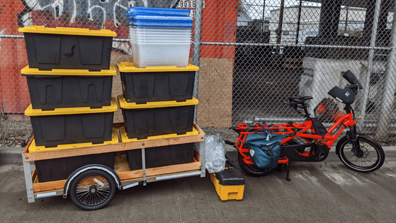 A red e-cargo bike sits parked to the right, with many boxes to the left, on the cargo flatbed area. A chainlink fence is in the background, with concrete below.