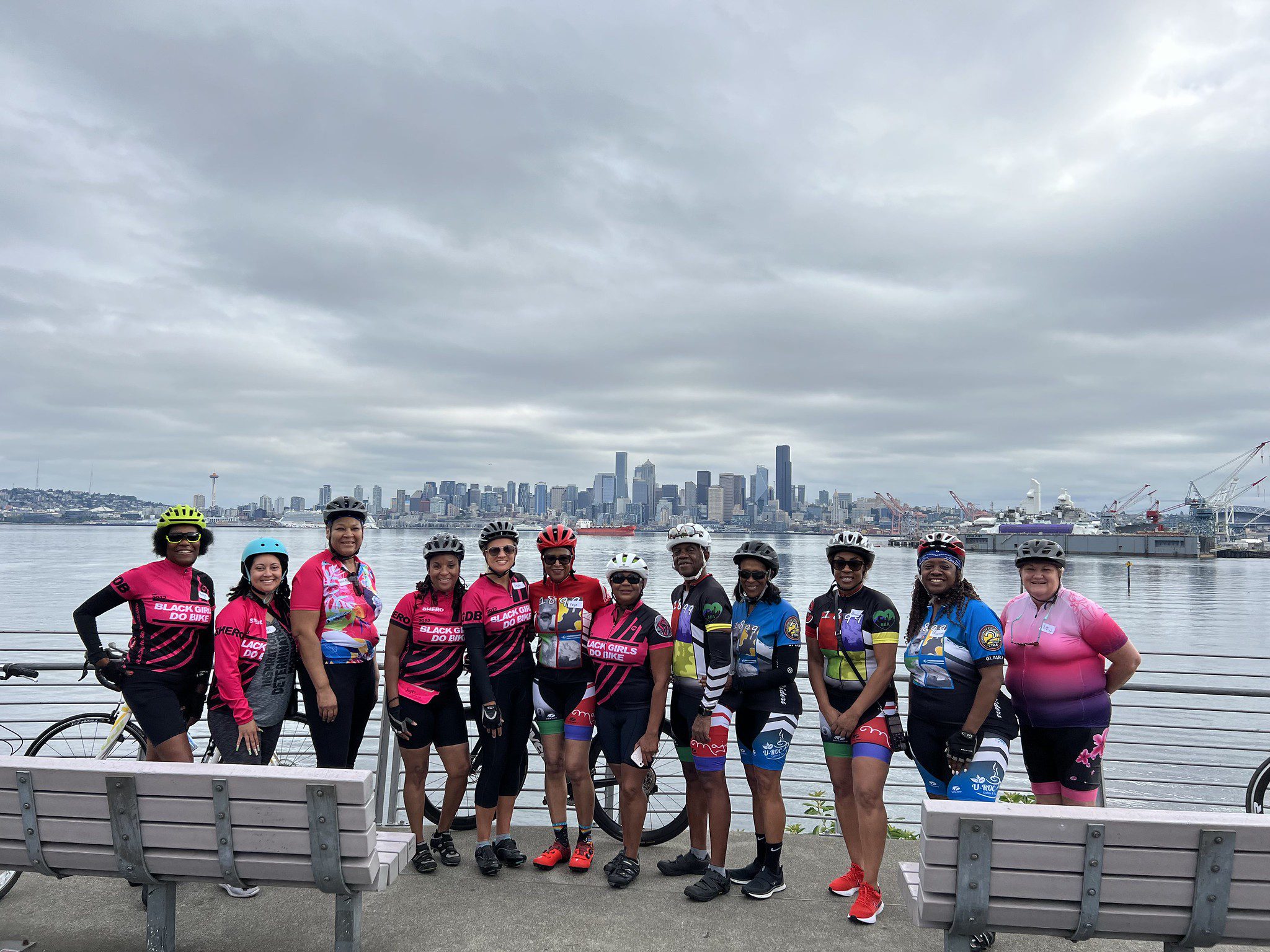 Photo of 12 people smiling while wearing biking outfits and helmets. The group is standing in front of water and a cityscape including large buildings in the background. Two benches are in the foreground.